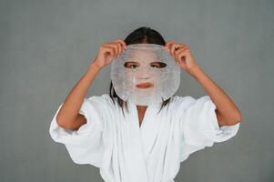 Against grey, background. Holding ,,mask for the face. Conception of beauty and self care. Young girl is in the studio photo