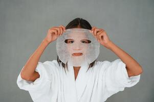 Against grey background,,. Holding mask for the face. Conception of beauty and self care. Young girl is in the studio photo