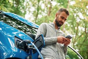 View from ,below,, with smartphone. Handsome man is standing outdoors near electric automobile and waiting for charging photo