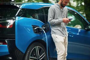 Wireless ,device, using ,smartphone. Handsome man is standing outdoors near electric automobile and waiting for charging photo