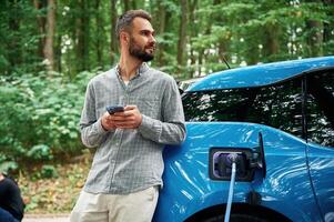 Trees behind.. Handsome ,,man is standing outdoors near electric automobile and waiting for charging photo