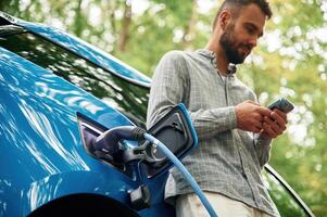 View, from below, with smartphone. Handsome man is standing outdoors near electric automobile and waiting for charging photo