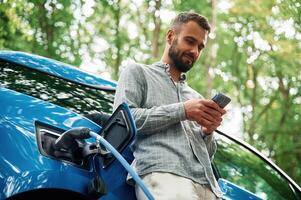 Trees, behind. Handsome man is standing outdoors near electric automobile and waiting for charging photo
