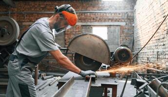 Using electrical saw to cut, the metal. Young factory worker in grey uniform photo