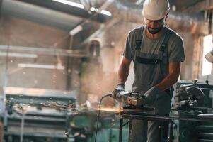 Angle ,grinder in hands. Young factory worker in grey uniform photo