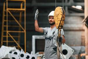 Using crane, hook. Young factory worker in grey uniform photo