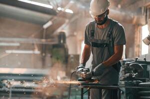 Angle, grinder in hands. Young factory worker in grey uniform photo