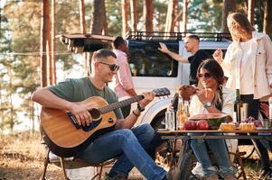Man is playing guitar, by table. Group of friends are together in the forest photo