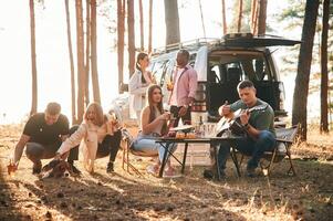 Drinking, eating, playing with dog... Group, of friends are together in the forest photo