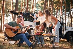 Man, is, playing guitar, by table. Group of friends are together in the forest photo
