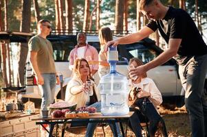 Sitting by the table with food, against car. Group of friends are together in the forest photo