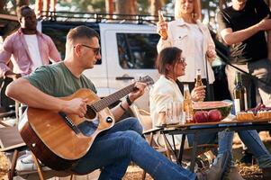 Man, is playing ,guitar, by table. Group of friends are together in the forest photo