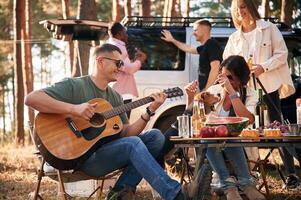 Man is playing guitar,, by, table. Group of friends are together in the forest photo