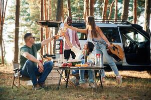Sittin,g by, the table with food, against car. Group of friends are together in the forest photo