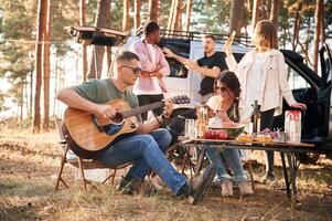 Man ,is ,playing guitar, by table. Group of friends are together in the forest photo