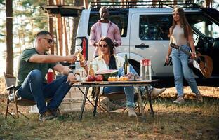 Sitting ,by the ,table with food, against car. Group of friends are together in the forest photo