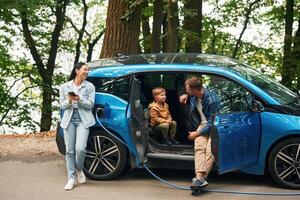 Road, in woodland. Mother,,, father and little son are waiting for electric car to charge outdoors photo
