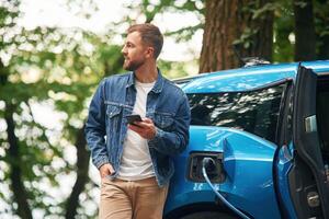 Standing and, holding smartphone.. Handsome man is waiting for his electric car to charge, outdoors photo