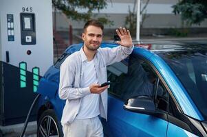 Standing with smartphone... Man with blue electric car on the charge station photo