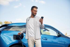 Standing with smartphone. Man with blue electric car on the charge station.. photo