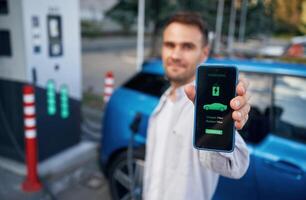 Charging process on the display of smartphone. Man with blue electric car on the station photo