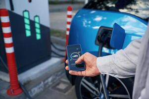 Showing process on the display, of, smartphone. Man with blue electric car on the charge station photo