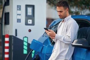 Side view, ,standing ,with smartphone, waiting. Man with blue electric car on the charge station photo