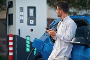 Side view,, standing with smartphone,, waiting. Man with blue electric car on the charge station photo