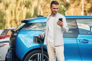 Wire in ,socket, holding ,smartphone.. Man with blue electric car on the charge station photo