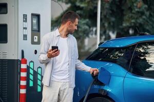 Wire, in socket, holding, smartphone... Man with blue electric car on the charge station photo
