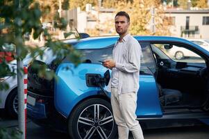 Front view. Man, with, blue electric, car, on the charge station photo