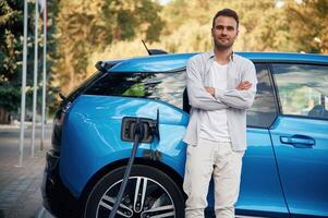 Standing with, arms ,crossed. Man with blue electric car on the charge station photo