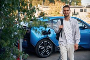 With backpack. Man, with, blue ,electric car on the charge station photo