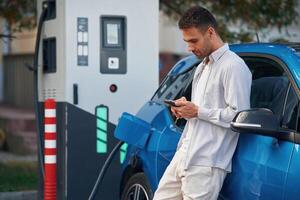 Side view, standing with smartphone,,, waiting. Man with blue electric car on the charge station photo