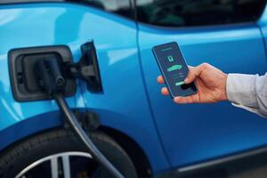 Charging, process on the display of smartphone. Man with blue electric car on the station photo