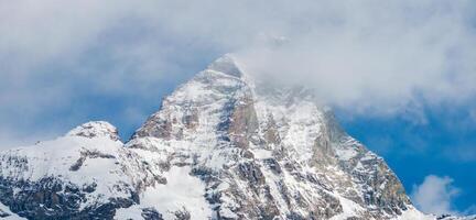 The Matterhorn mountain in the Alps features rugged, snow covered slopes, a triangular peak, and wisps of clouds against a clear blue sky. photo