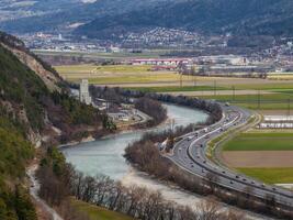 View of the Austrian Alps showing a river, a modern highway with traffic, a castle like stone tower, and a small town with fields and buildings. photo