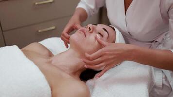Close up side view beautiful young woman lying on bed at beauty and spa salon enjoying lymphatic facial massage which removes excessive fluid from face, decreases puffiness and produces lifting effect video