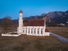 St. Coloman Church stands in an open field with bare trees, framed by the Bavarian Alps and a clear blue sky. Snow capped peaks are visible in the distance. photo
