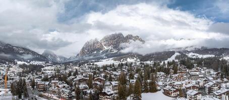 Aerial view of Cortina d'Ampezzo in Italy with snow covered rooftops, evergreen trees, Dolomites peaks, low clouds, and a visible construction crane. photo