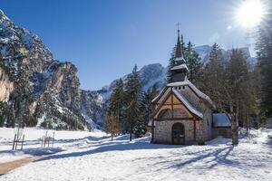 A small chapel with a pointed roof and cross stands amid snow covered ground, evergreen trees, and rugged mountain cliffs at Braies Lake in Italy. photo