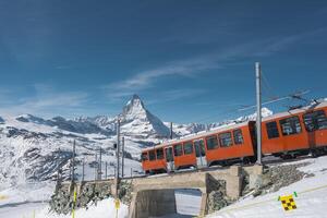 A red train crosses a small bridge in snowy Zermatt, Switzerland, with the Matterhorn mountain and clear blue skies creating a striking alpine scene. photo