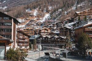 Zermatt in Switzerland features wooden chalets, modern buildings, a river with a pedestrian bridge, and snow dusted alpine slopes with scattered trees. photo