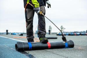 A worker in protective gear uses a propane torch to apply black roofing membrane on a flat rooftop, with additional materials and cloudy sky visible. photo