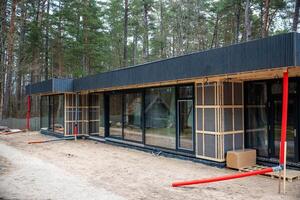 A modern single story house with black exterior and large glass windows under construction, featuring red ventilation pipes and surrounded by tall pine trees. photo