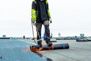 A worker in protective gear uses a torch to apply roofing material on a flat rooftop. Rolls of material and distant structures are visible under a cloudy sky. photo