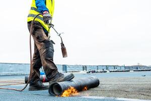 A worker in a high visibility vest uses a torch to heat and apply roofing material on a flat rooftop. Tools and rolls of material are visible under a cloudy sky. photo