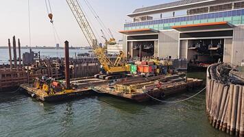 New York. US 05.04.2025 Construction barge with crane operating at ferry terminal on sunny day in New York harbor, Workers and equipment positioned near industrial pier during dock maintenance work video