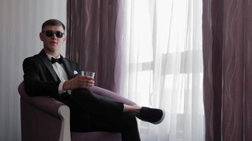 Stylish man in tuxedo and sunglasses holding drink while sitting in hotel room armchair, Modern groom or guest relaxes in daylight with confident pose near window curtains video
