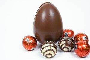 A large chocolate egg and several chocolates are placed on a white background. Three round candies are wrapped in red foil, and the other two have a chocolate striped shell, creating a sweet treat. photo